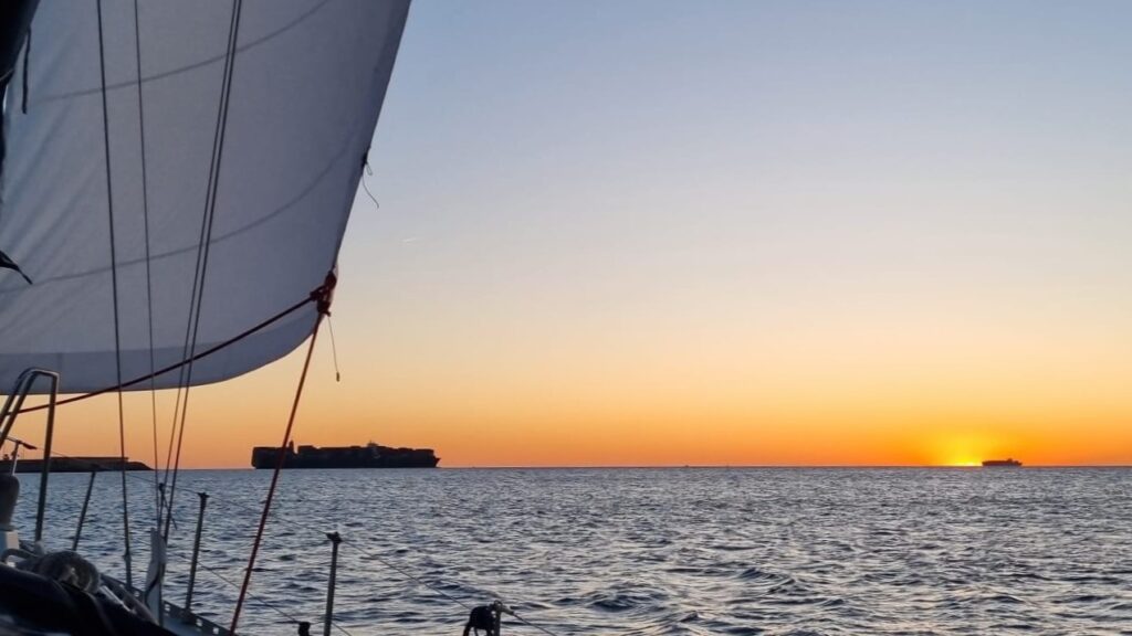 El sol se asoma en el horizonte con un buque mercante que casi lo tapa. En primer plano, el Atlantis navega rumbo al puerto de Valencia.