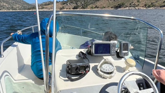 Un tripulante maneja con su mano derecha el motor de la lancha Avante con radio y aparatos electrónicos delante. En proa un alumno se protege del viento con capucha azul